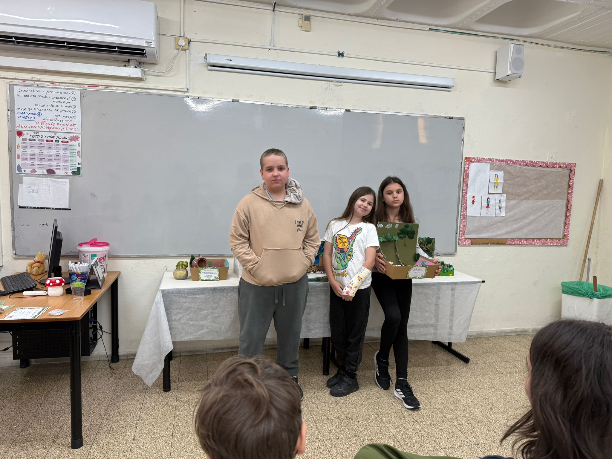 צילום מהכיתה: תלמידים מציגים דגמים שהכינו, לצד עבודות נוספות על שולחן